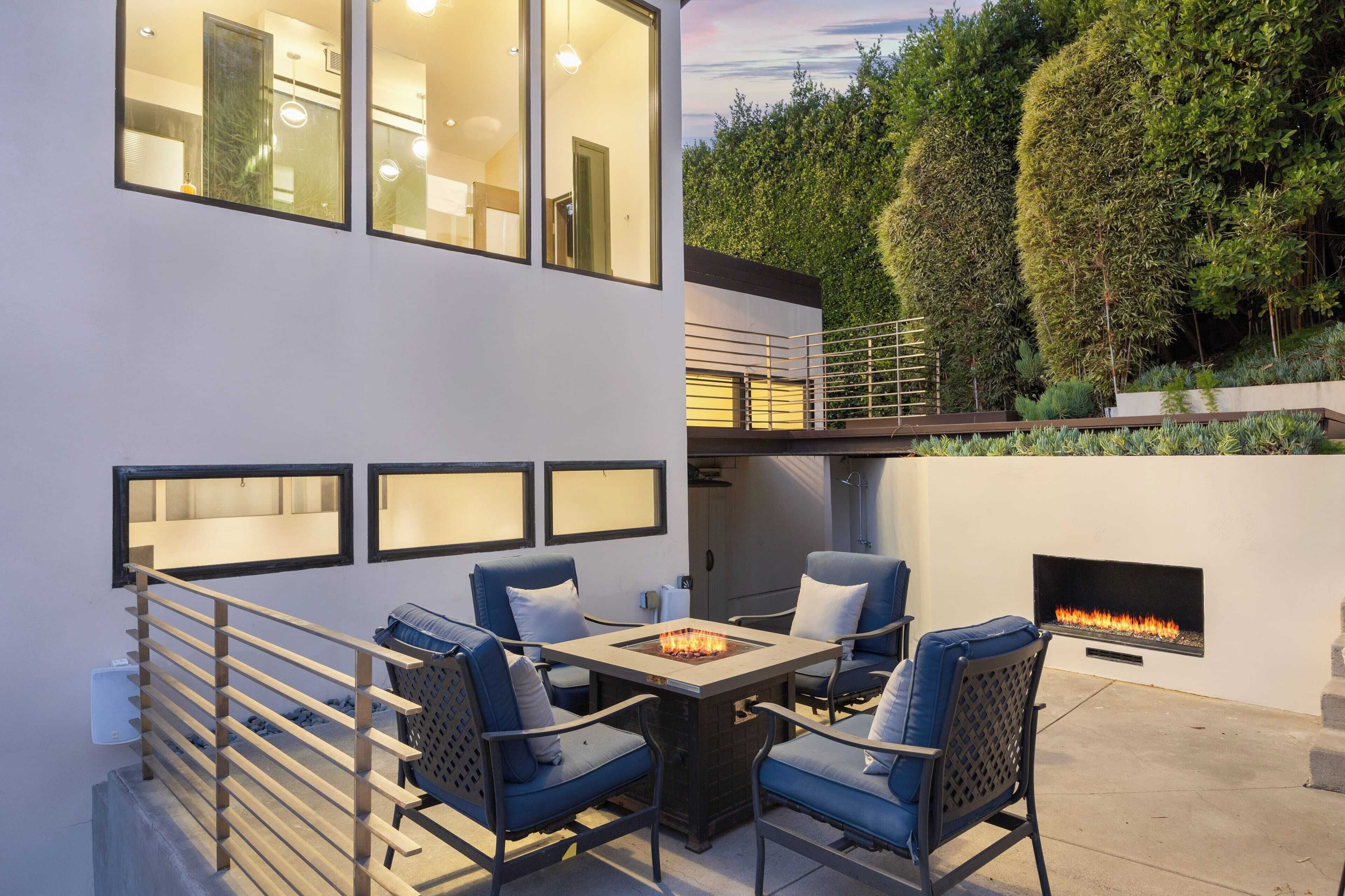 An outdoor patio with a fire pit table and chairs, surrounded by a white wall with large windows and a view of a hedge.