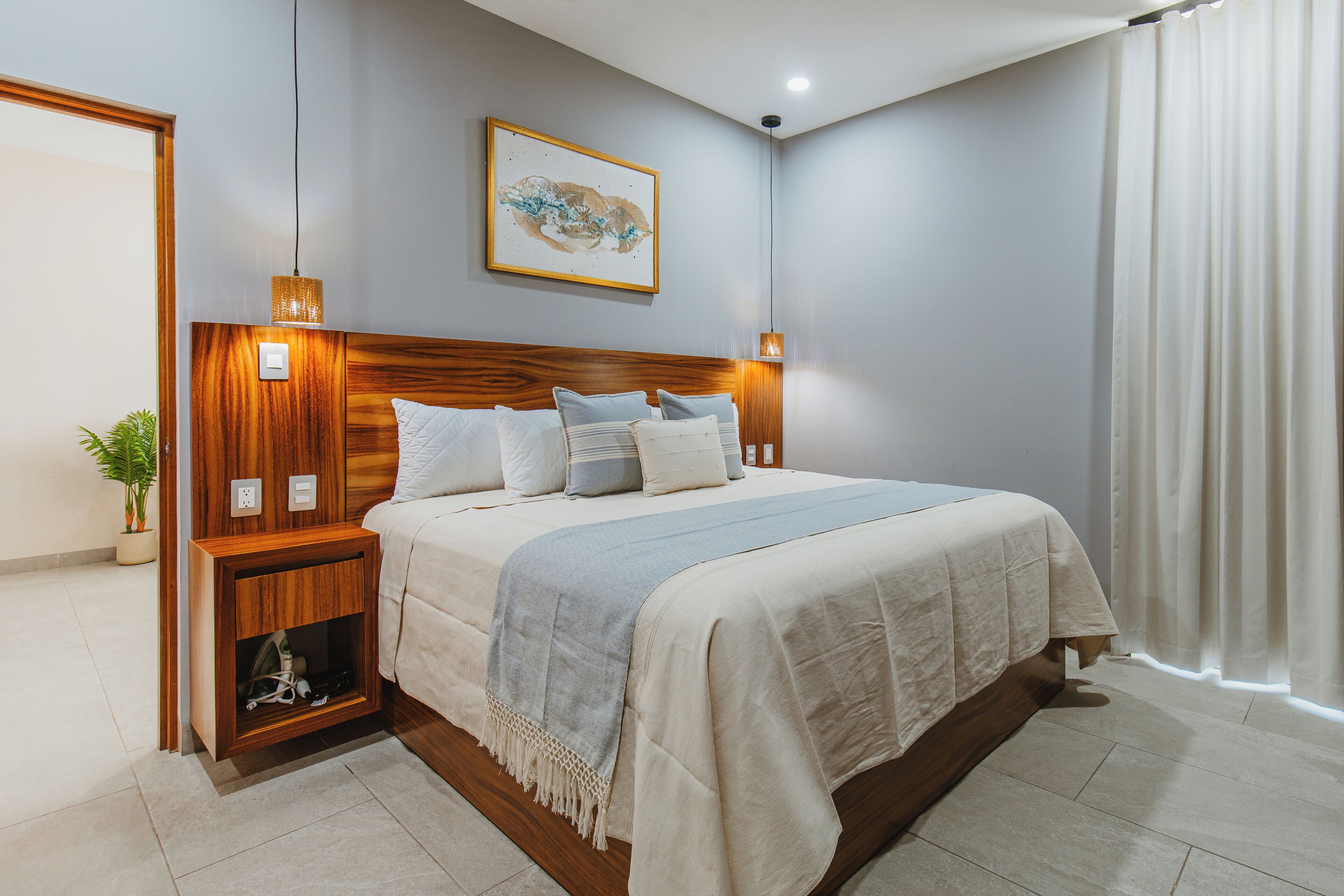 A modern bedroom with a wooden headboard, a large bed with multiple pillows, a bedside table with a lamp, and a wall-mounted artwork.