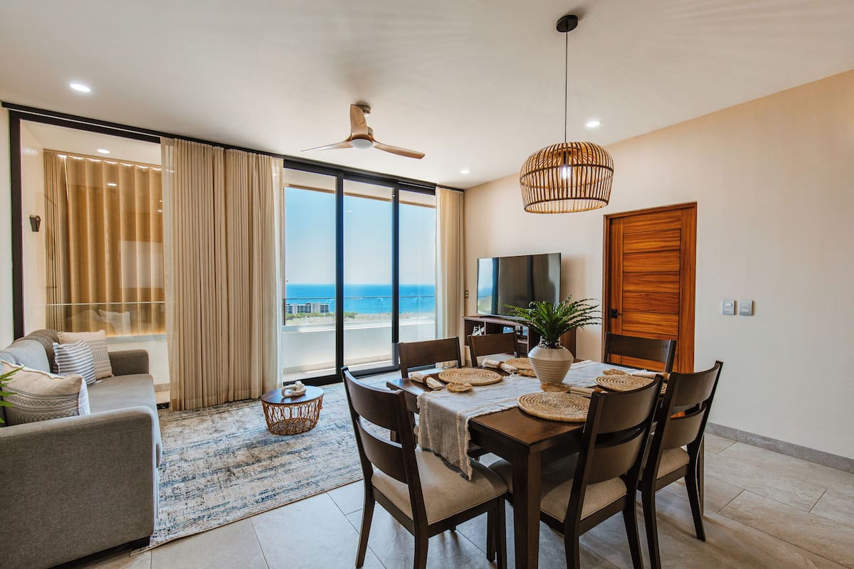 A dining area with a table set for four, a ceiling fan, and a view of the ocean through large windows.