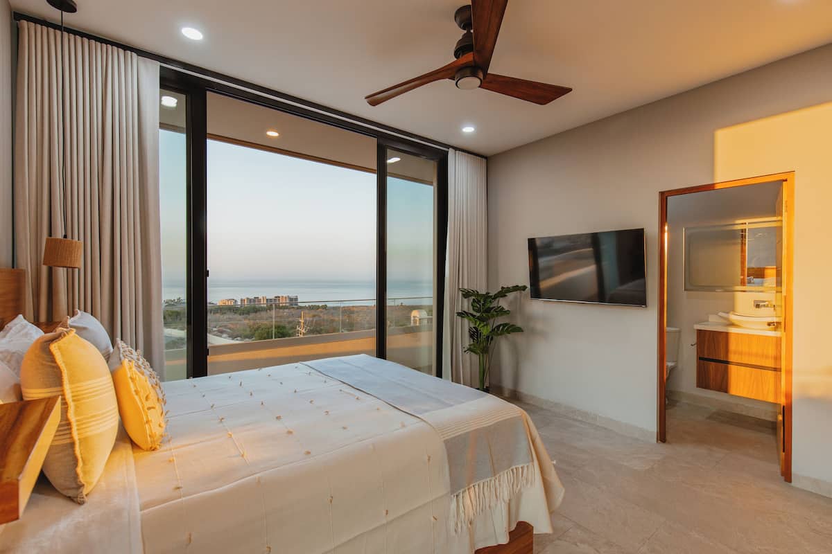 A bedroom with a large bed, a ceiling fan, a TV, and a view of the city.