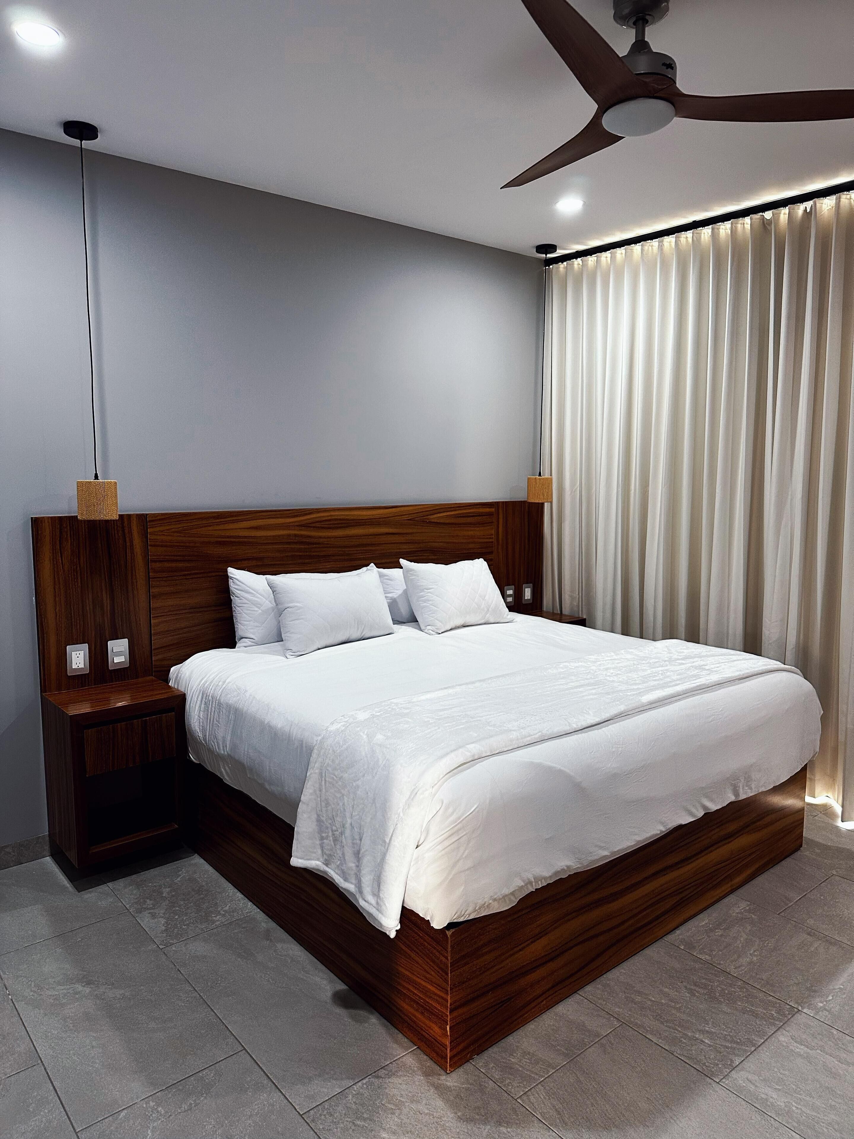 A modern bedroom with a wooden headboard, a large bed with white bedding, a nightstand, a wall-mounted lamp, and a ceiling fan.
