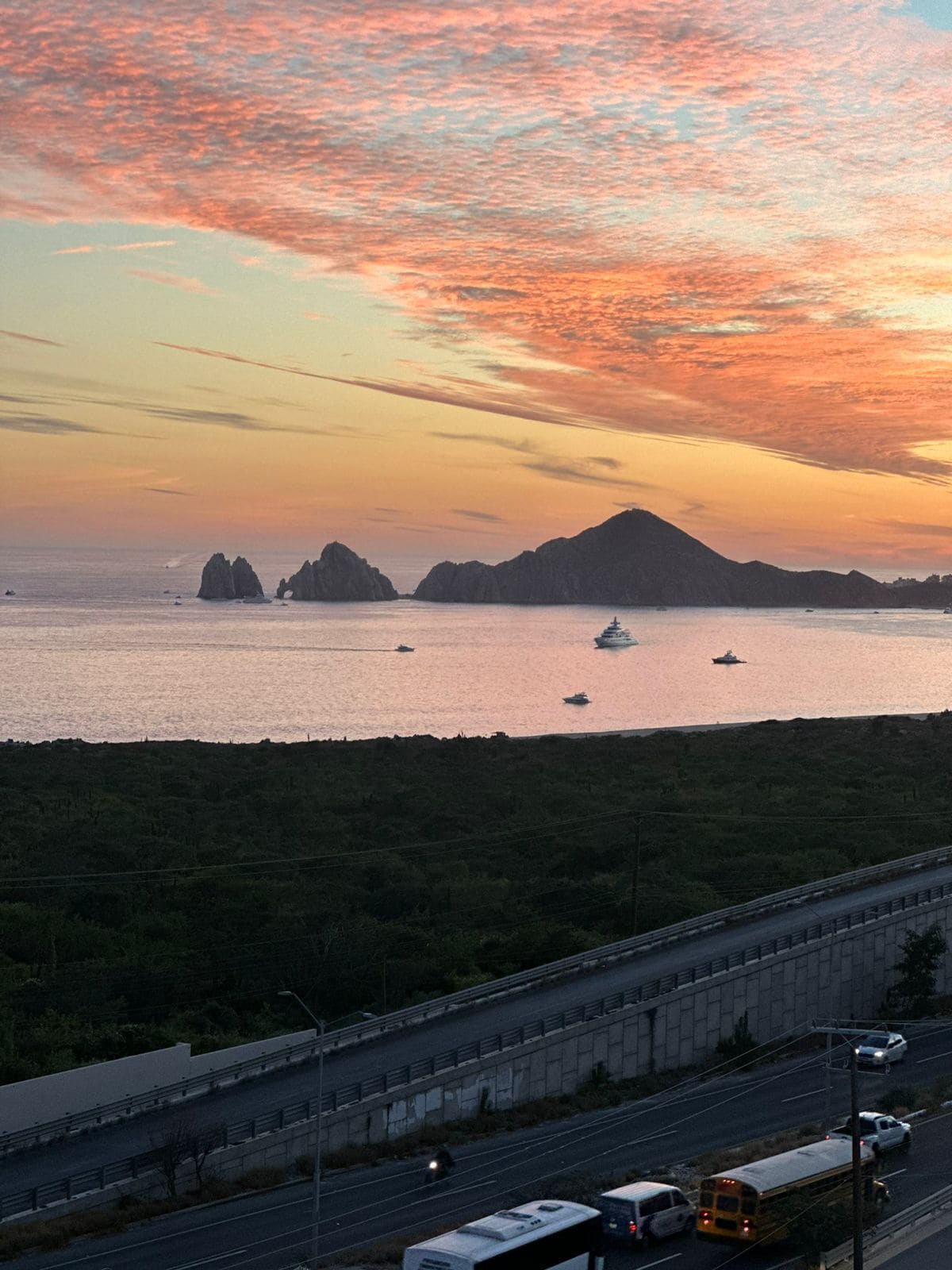 Une route côtière offrant une vue sur le coucher de soleil, avec un pont, des véhicules et des îles au loin.