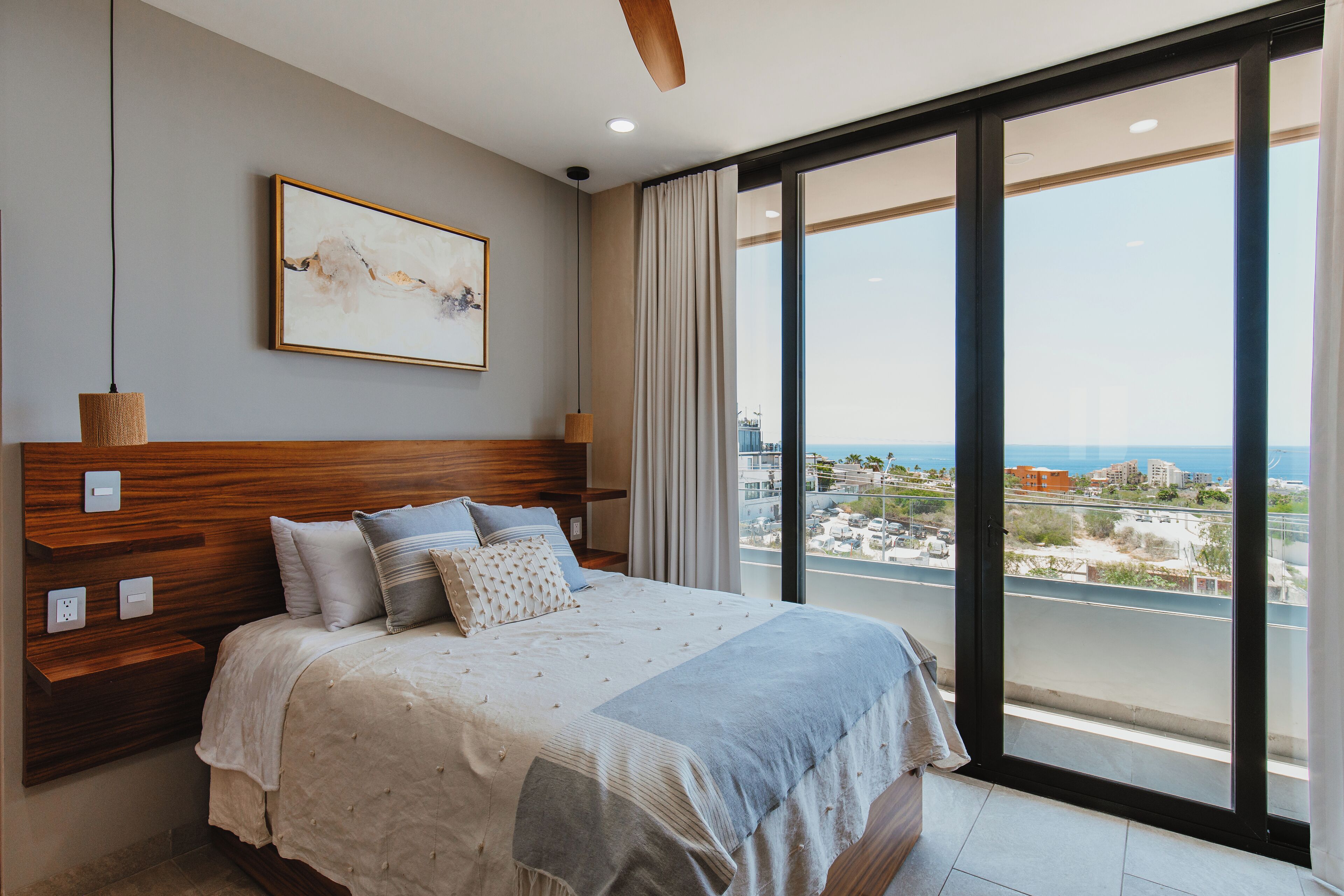 Ein Schlafzimmer mit einem großen Bett, einem Holz-Kopfteil, Blick auf die Stadt durch große Fenster und einem Deckenventilator.