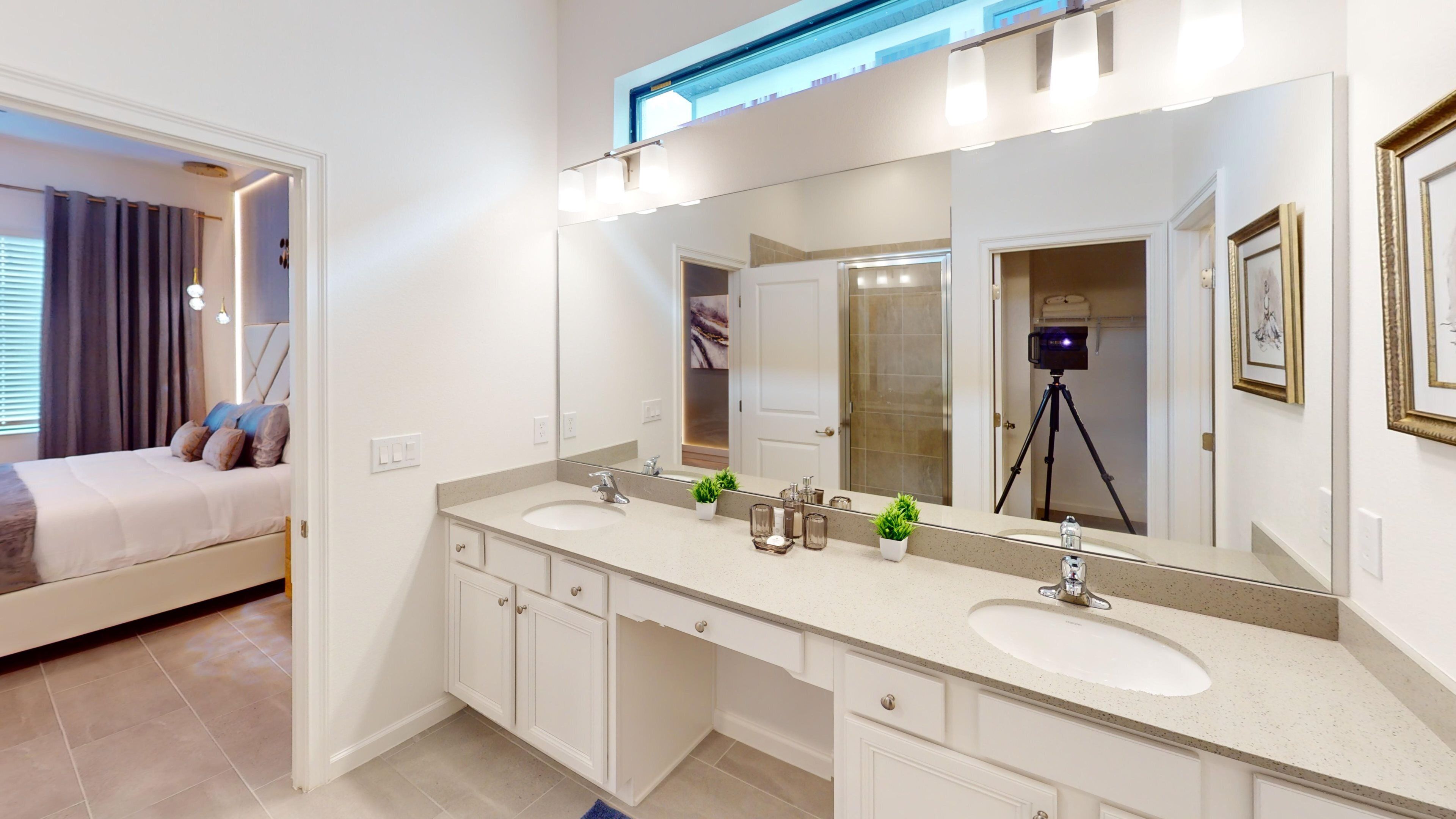 A modern bathroom with two sinks, a large mirror, and a bed in the adjacent room.