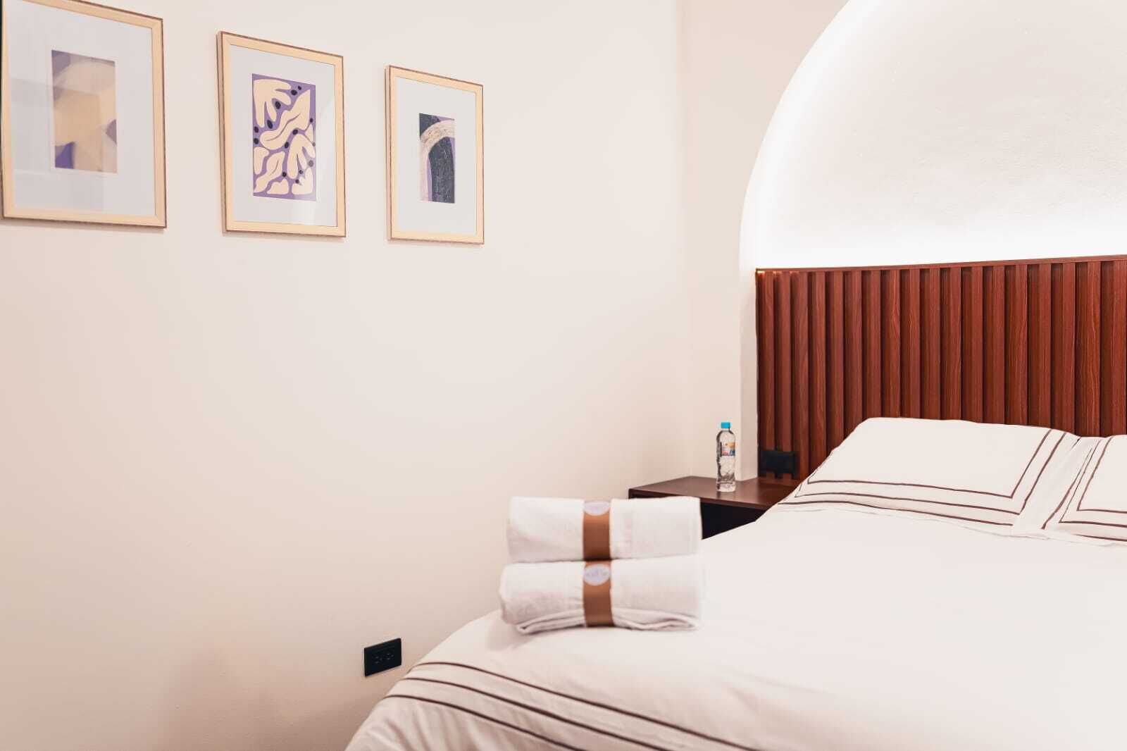 A hotel room with a neatly made bed, a nightstand with a bottle, and three framed artworks on the wall.
