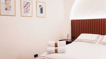 A hotel room with a neatly made bed, a nightstand with a bottle, and three framed artworks on the wall.