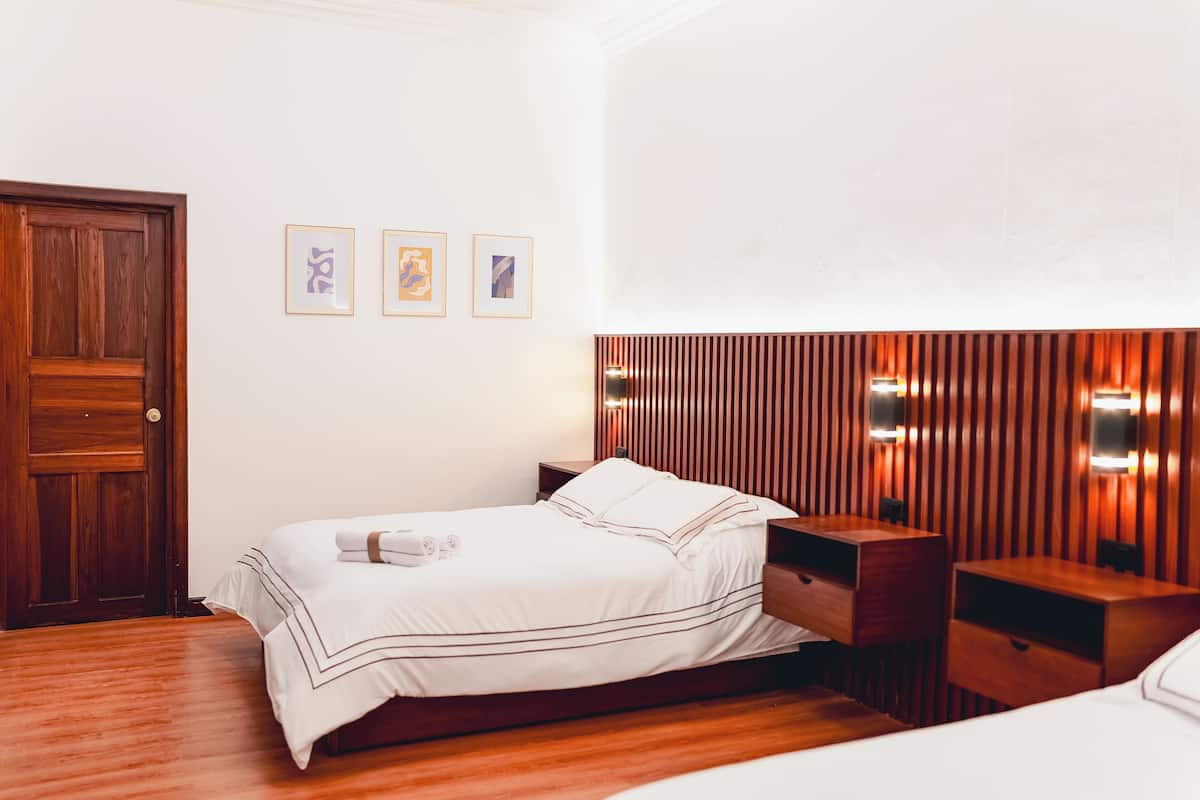 A hotel room with a wooden door, a bed with white bedding, a wooden headboard with built-in lighting, and three framed pictures on the wall.