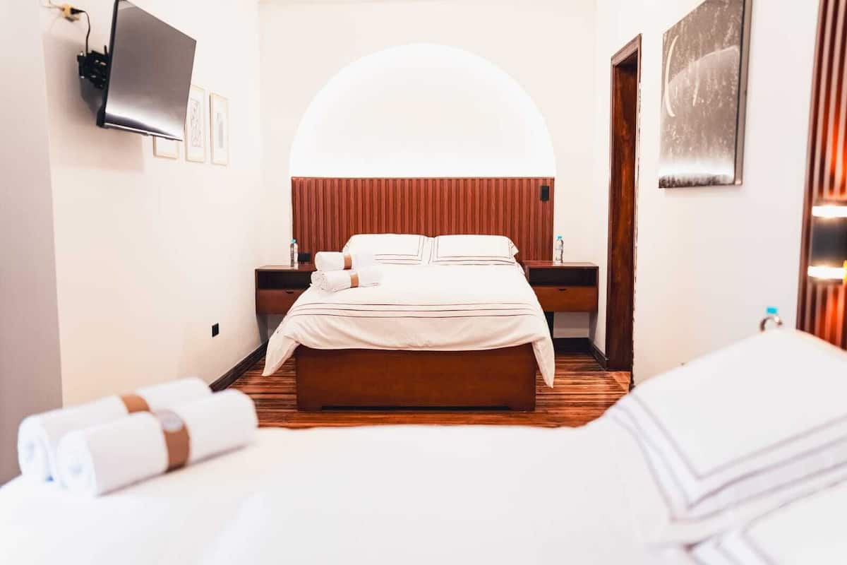 A hotel room with a bed, a TV mounted on the wall, a wooden floor, and a large arched window.