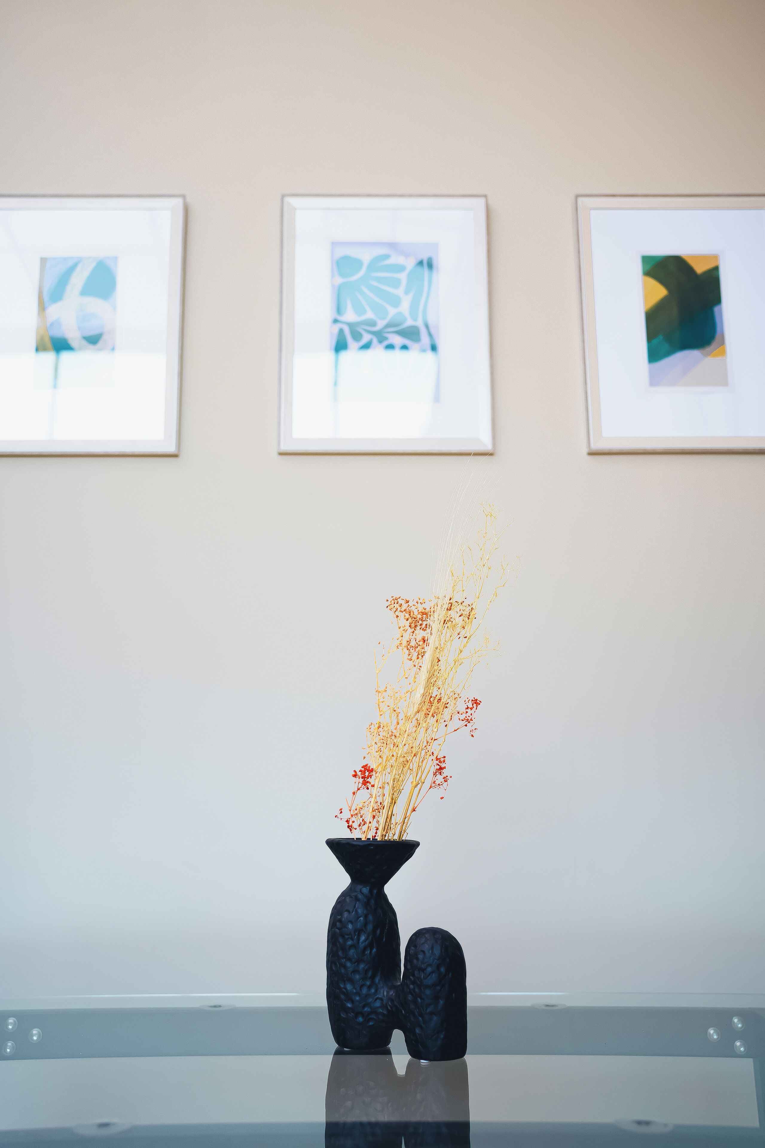 Una habitación con una mesa de vidrio, un florero con flores secas y tres obras de arte enmarcadas en la pared.