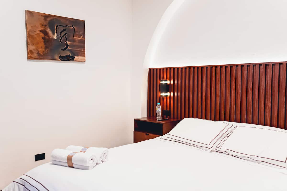 A neatly made bed with white linens, a wooden headboard, a nightstand with a bottle and a lamp, and a wall-mounted artwork.