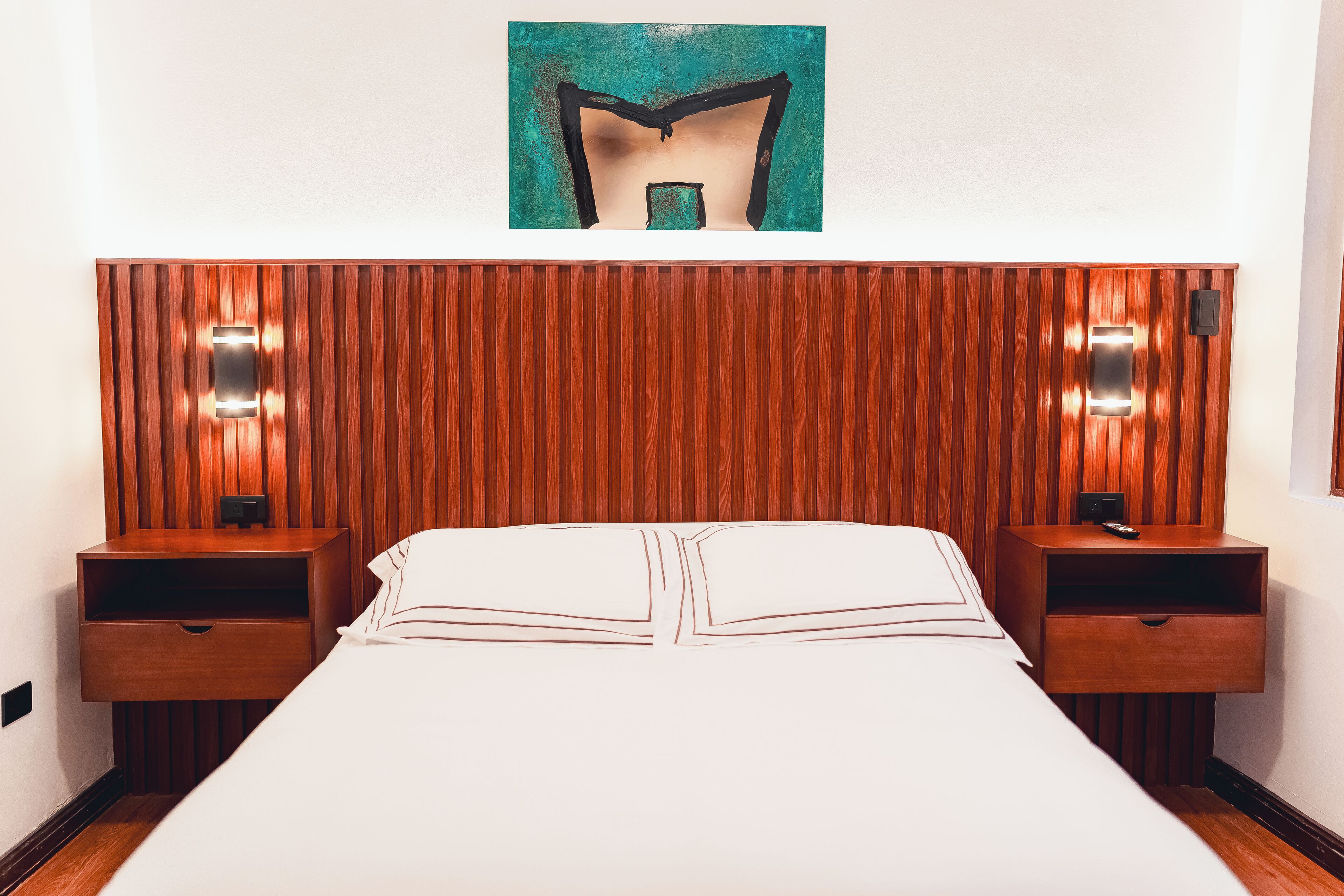 Habitación de hotel con cabecera de madera, dos mesitas de noche con lámparas, una cama grande con ropa de cama blanca y una obra enmarcada sobre la cabecera.