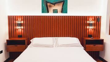 Habitación de hotel con cabecera de madera, dos mesitas de noche con lámparas, una cama grande con ropa de cama blanca y una obra enmarcada sobre la cabecera.
