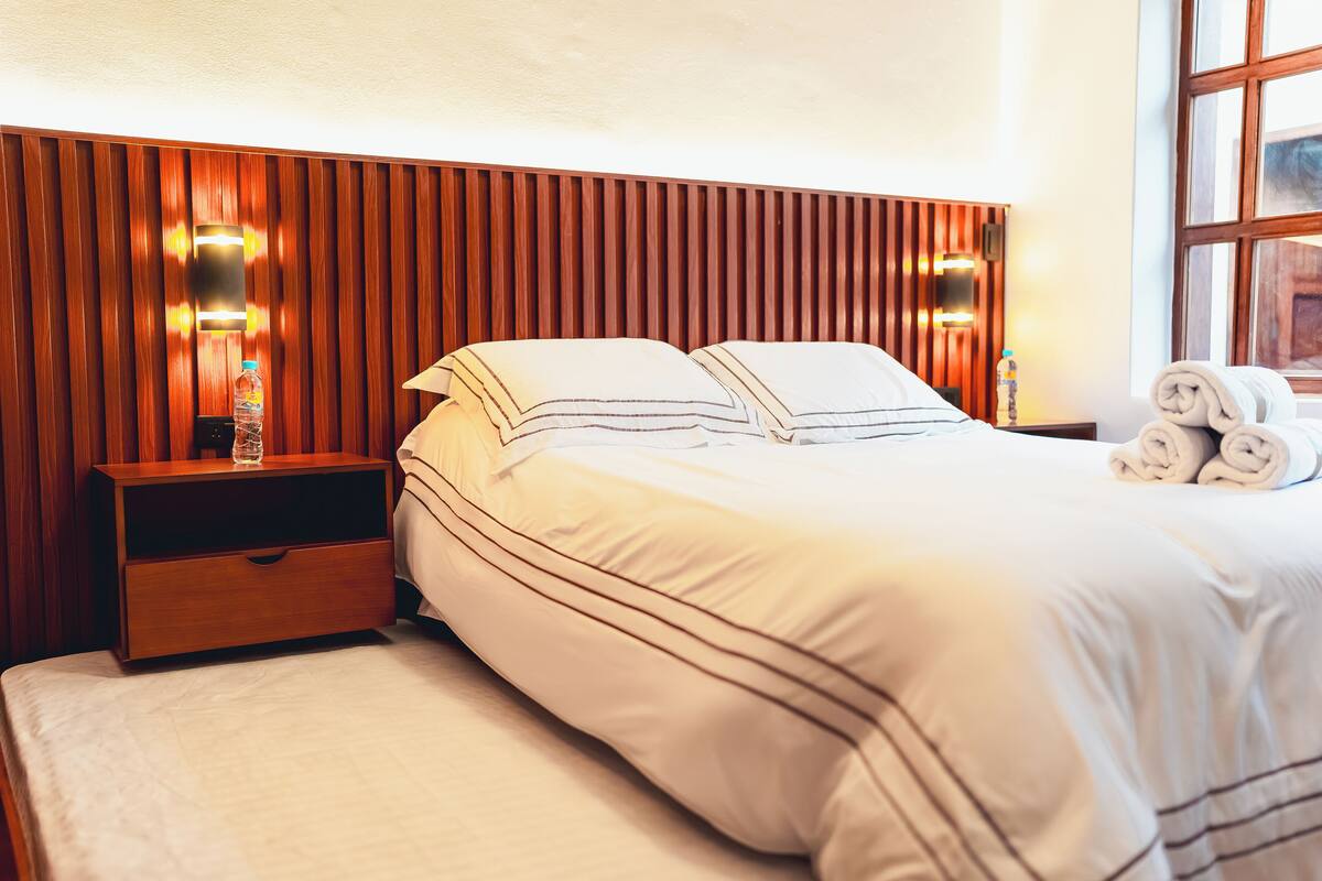 A hotel room with a wooden headboard, a bed with white bedding, a nightstand with a bottle and a lamp, and a window with rolled towels on the bed.