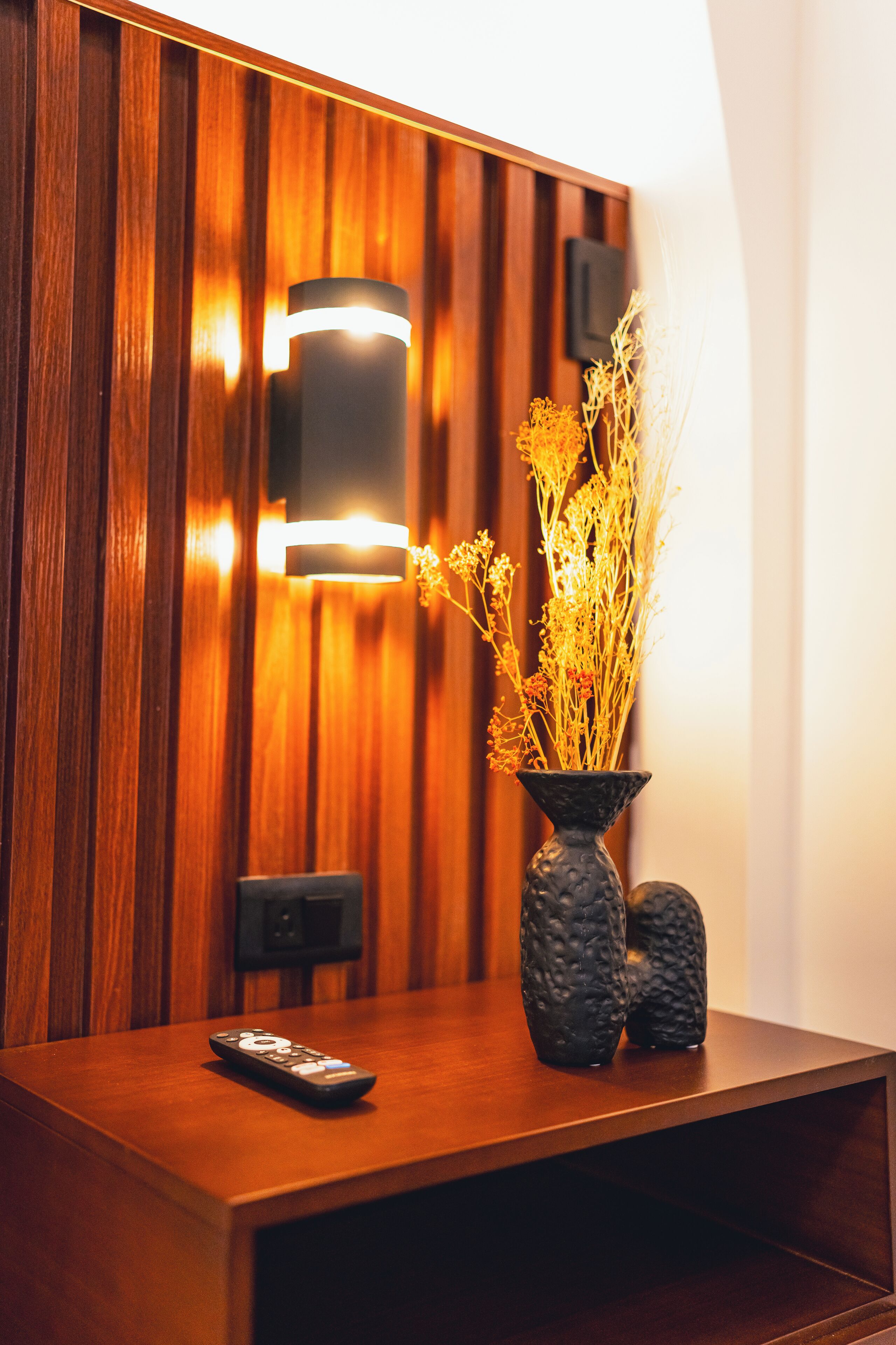 A wooden reception desk with a vase of dried flowers, a remote control, and a wall-mounted light fixture.