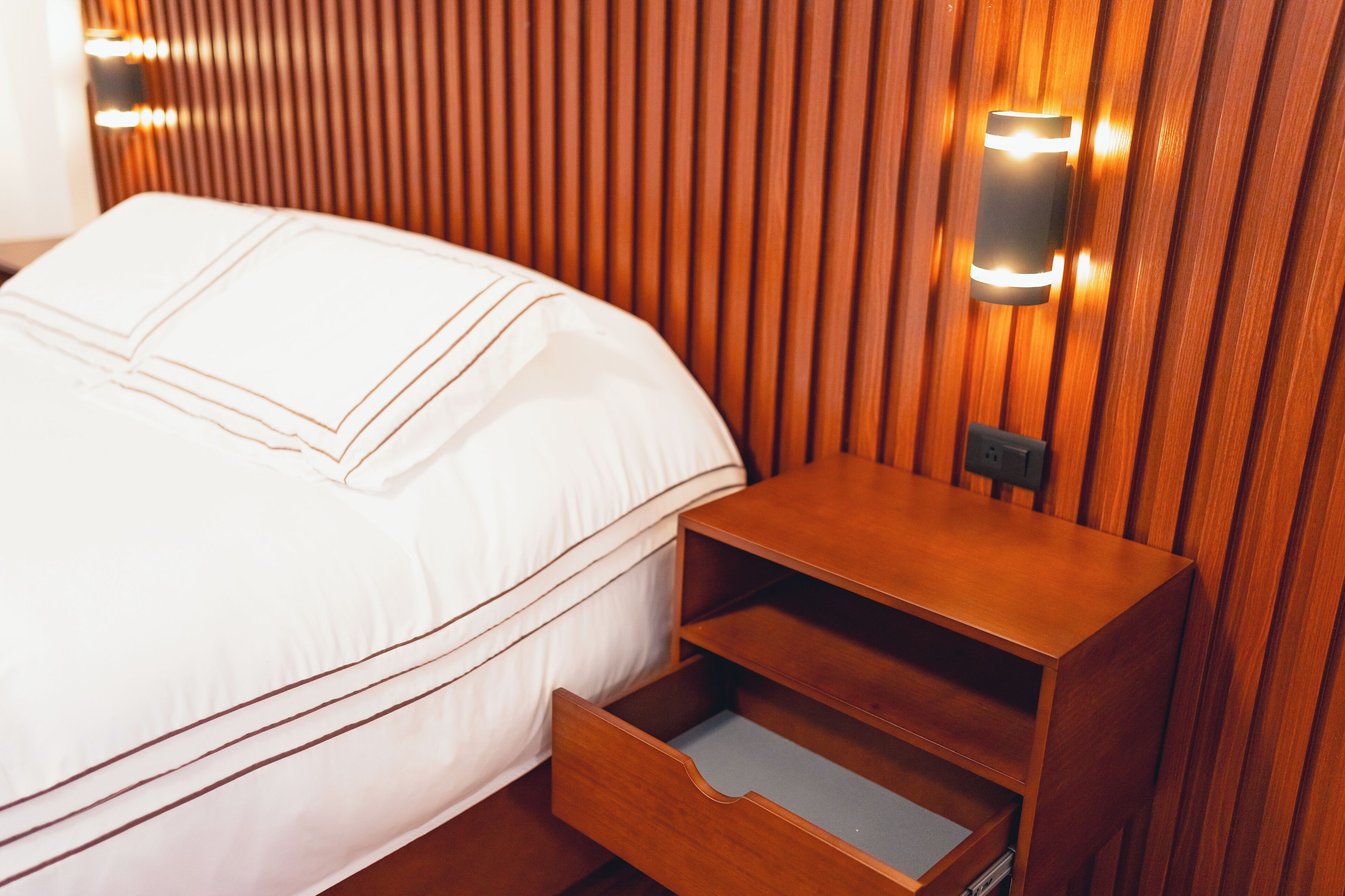 Una cama bien hecha con sábanas blancas y una mesita de noche de madera con un cajón.