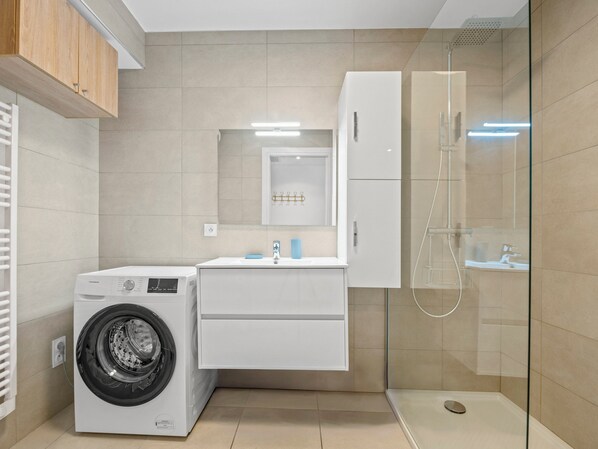 Un moderno cuarto de lavado con lavadora de carga frontal, encimera con lavabo y espejo, y cabina de ducha con mampara de vidrio.