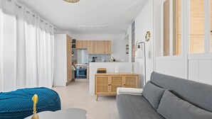 Ein modernes Wohnzimmer mit einem grauen Sofa, einem blauen Sessel und einem kleinen Tisch mit einem dekorativen Gegenstand. Im Hintergrund ist eine Küche mit Holzschränken und einem eingebauten Ofen zu sehen.