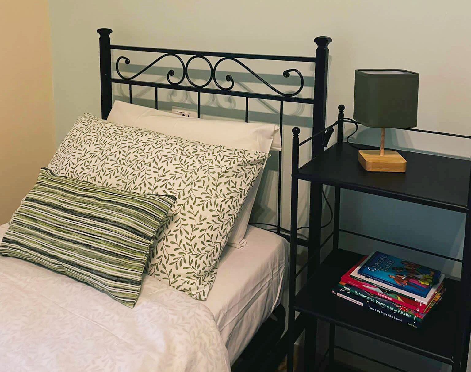 A neatly made bed with patterned pillows, a bedside table with a lamp, and a stack of books.