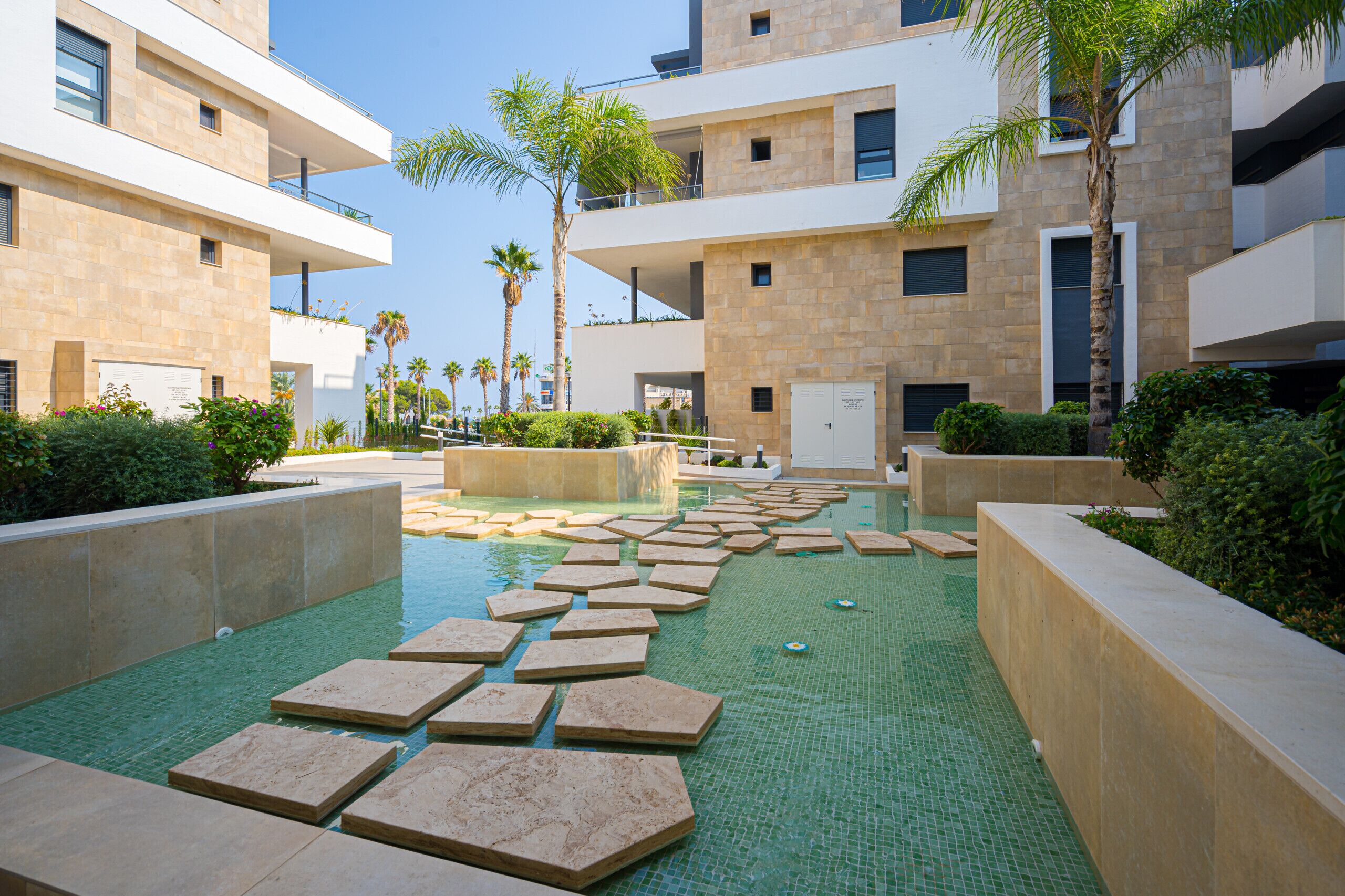 A modern building with multiple balconies, a pool with stepping stones, and surrounding greenery.