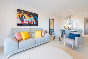 A modern living room with a grey sofa, a dining area with a table and chairs, and a large abstract painting on the wall. - Apartamento Flamenca Village (Orihuela Costa)