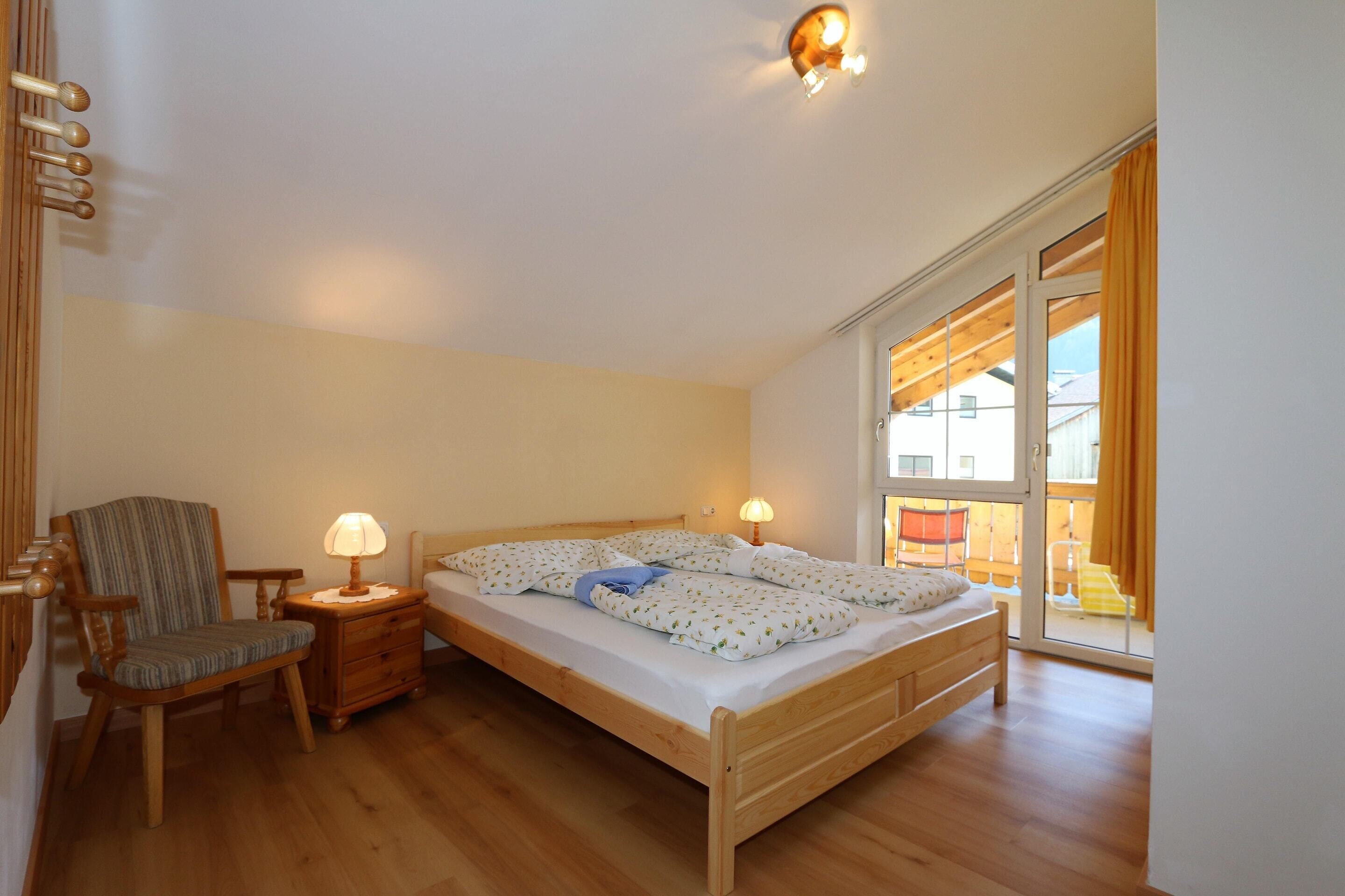 Ein Schlafzimmer mit einem hölzernen Bett, einem Sessel, einem Nachttisch mit Lampe und einem großen Fenster mit Vorhängen.