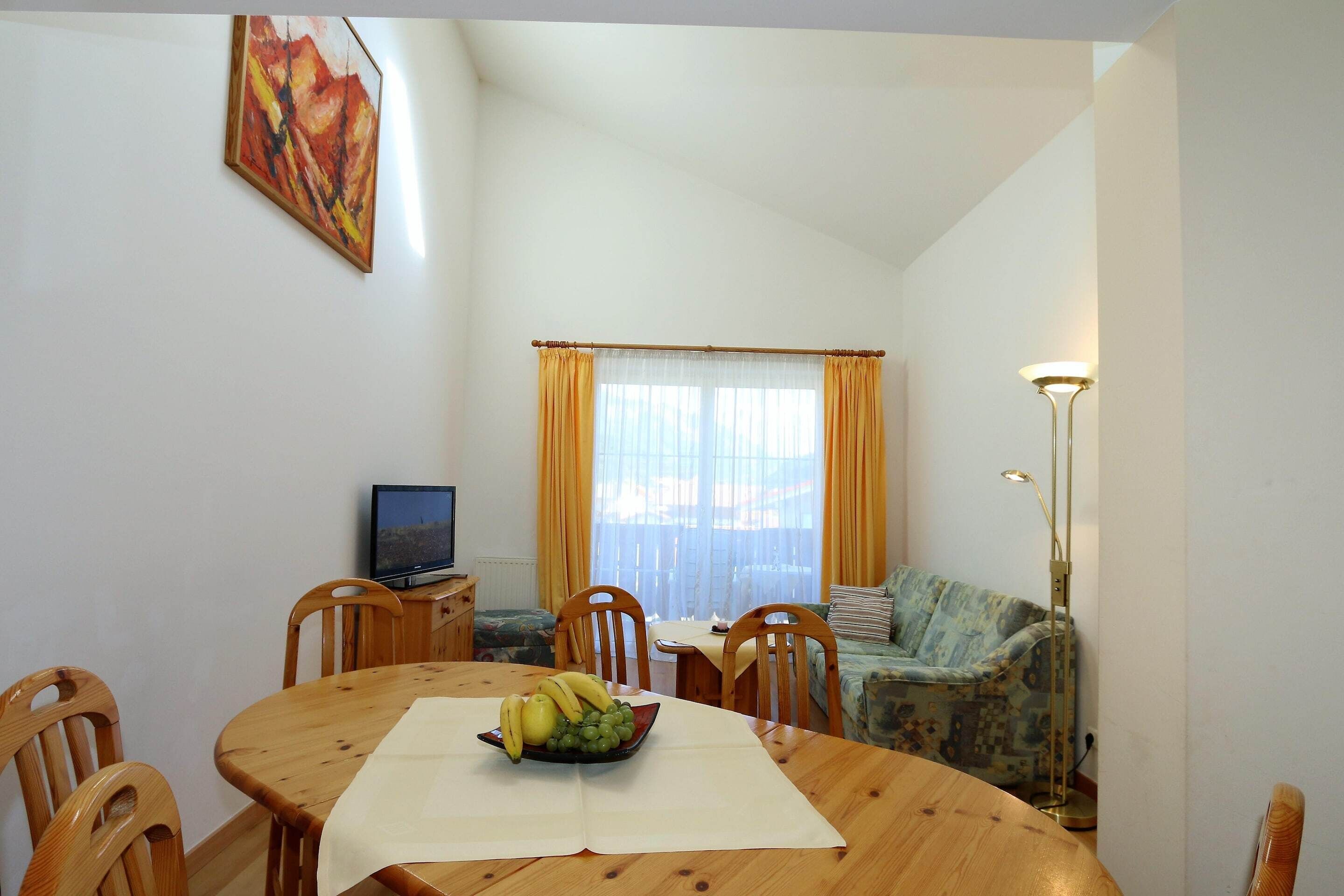 A dining area with a wooden table, chairs, a fruit bowl, a television, and a sofa.