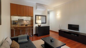 Un soggiorno moderno con un divano, un tavolino e una TV su un mobile nero. È presente una zona cucina con mobili in legno e un forno a microonde.
