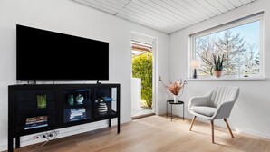 A modern living room with a flat-screen TV, a bookshelf, a grey armchair, and a window with a view of trees.