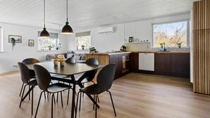 A modern kitchen with a dining area, featuring a wooden floor, pendant lights, and a view of the outdoors through large windows.