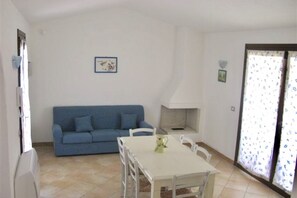A room with a blue sofa, a white table with chairs, a white fireplace, and a window with patterned curtains.