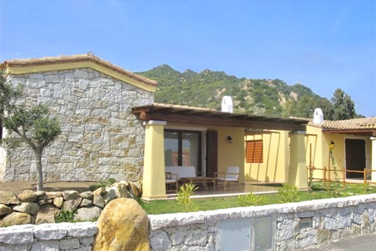 A stone building with a yellow facade, a patio with outdoor seating, and a mountainous backdrop.