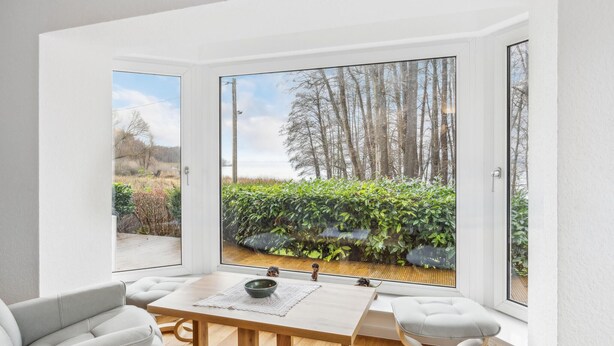 Zona pranzo moderna con tavolo e sedie in legno, ampie finestre con vista su giardino e alberi.