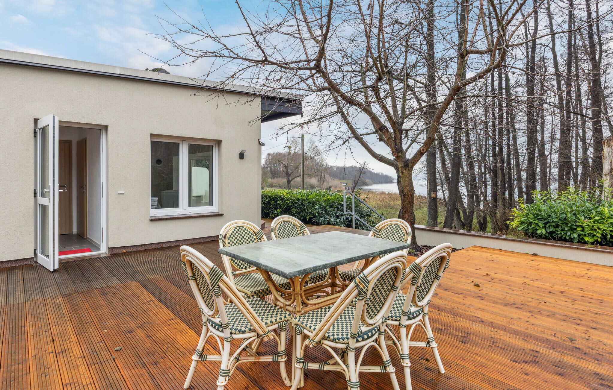 Eine Terrasse im Freien mit Tisch und Korbstühlen, einer Tür sowie Blick auf Bäume und ein Gewässer.