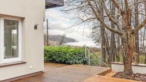 Un pontile in legno conduce a una casa con un'ampia finestra, vista su un lago e un albero sullo sfondo.