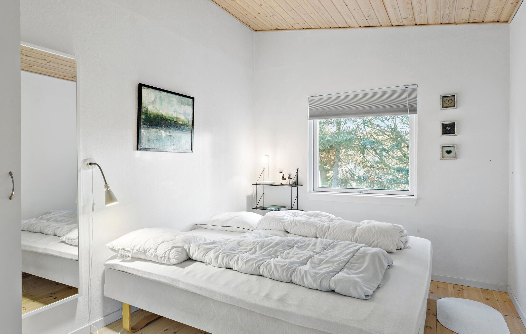 Een minimalistische slaapkamer met een bed, een nachtkastje met een lamp, een raam met uitzicht op bomen en een lijst met een afbeelding aan de muur.