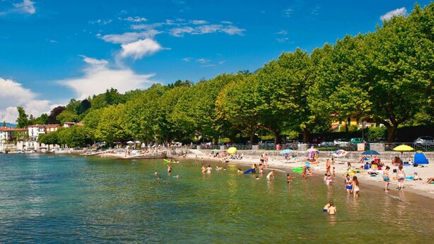 A lakeside beach with clear water, boats, and a forested hillside.