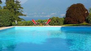 A clear blue swimming pool with two red lounge chairs facing the water, surrounded by greenery and a view of a distant coastline.