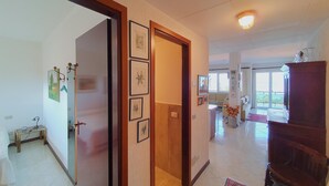 A spacious hallway with a wooden door, framed artwork on the walls, and a view into a living area with a sofa and a dining table.
