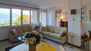 A living room with a sofa, a coffee table, and a vase with flowers. There are windows with a view of trees and a lamp on a side table.
