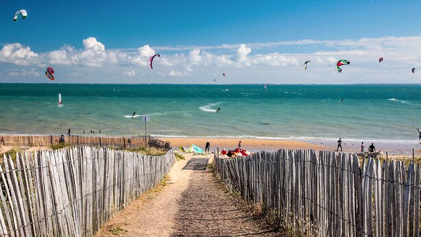 Una spiaggia con una recinzione di legno, cielo sereno e diversi aquiloni in volo.