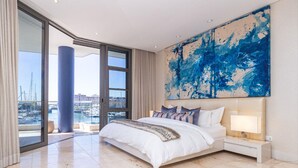 A spacious bedroom with a large bed, a nightstand, and a view of a marina through floor-to-ceiling windows.