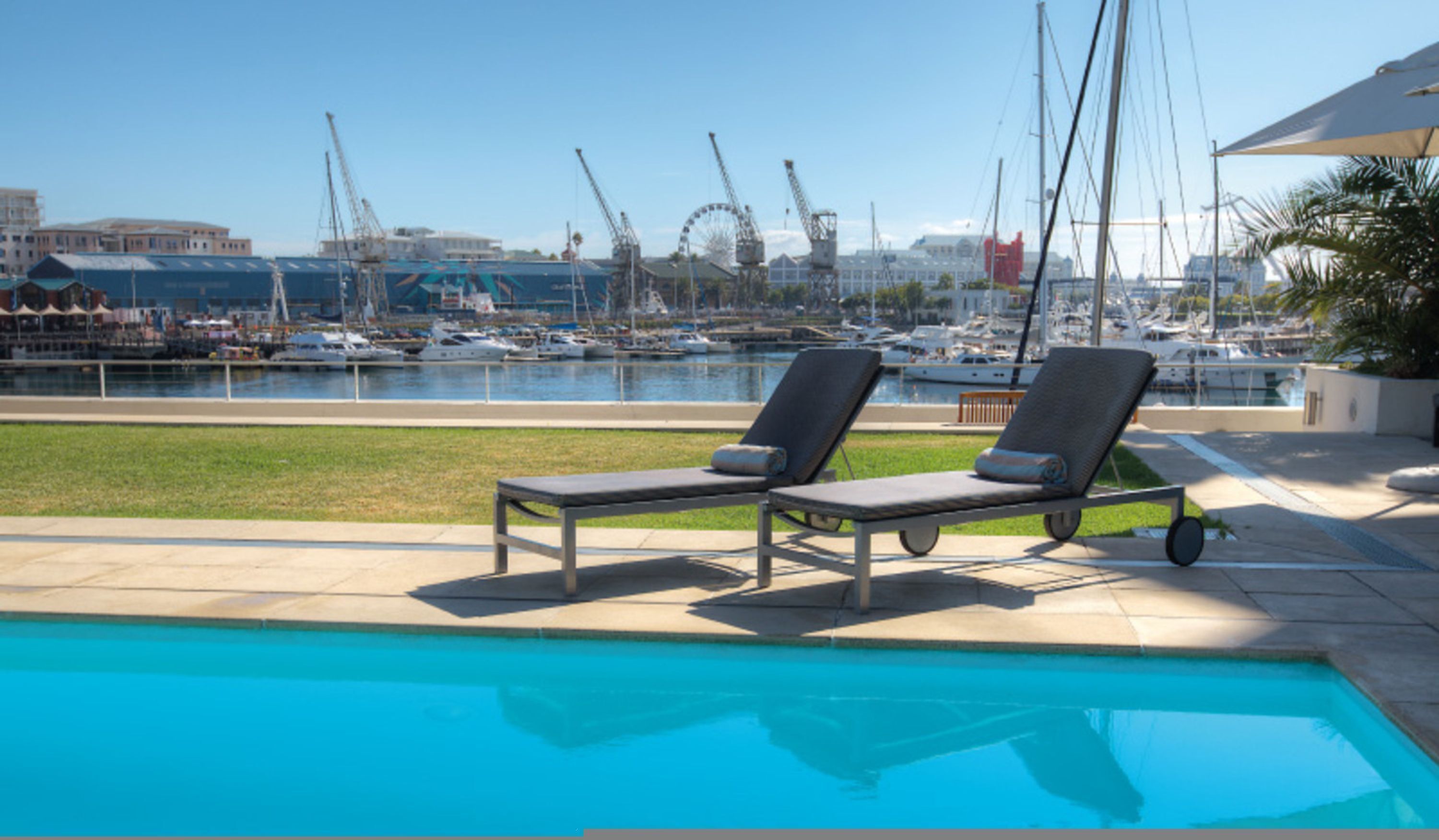 Una piscina con dos sillas reclinables, un puerto deportivo con barcos y una noria al fondo.