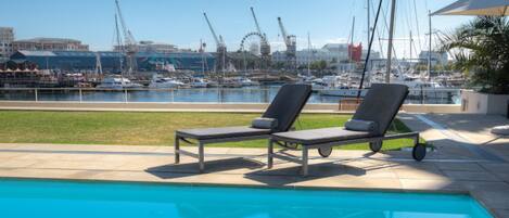 Una piscina con dos sillas reclinables, un puerto deportivo con barcos y una noria al fondo.