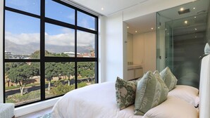 Un dormitorio moderno con un ventanal que ofrece vistas a la ciudad, una cama bien hecha con almohadones estampados y un divisor de ambiente de vidrio.