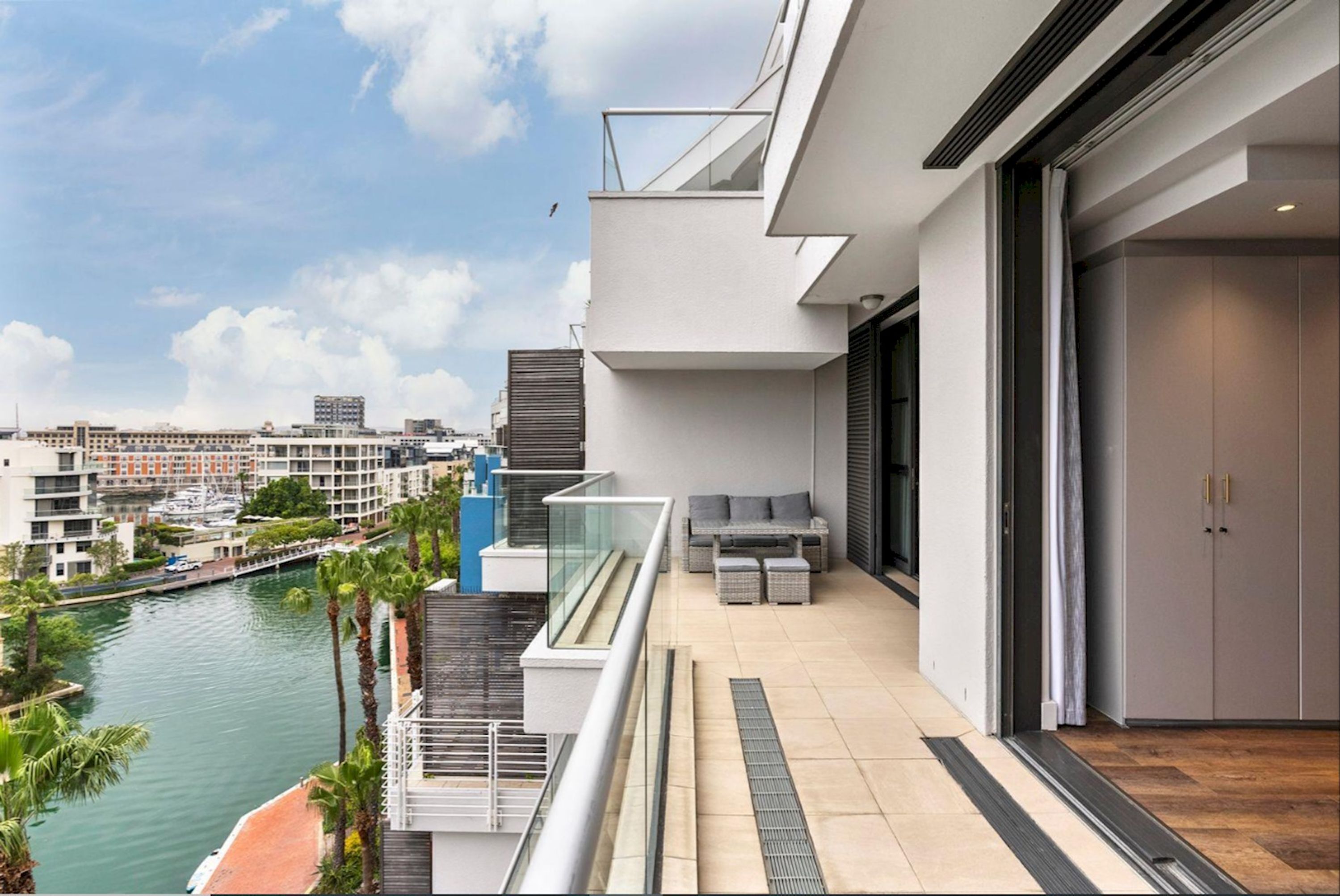 A modern balcony with a view of a river and cityscape, featuring a glass railing, outdoor seating, and a sliding door.