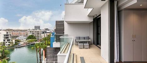 A modern balcony with a view of a river and cityscape, featuring a glass railing, outdoor seating, and a sliding door.