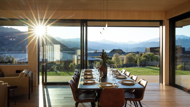 Un comedor con una mesa larga dispuesta para una comida, amplios ventanales que ofrecen vistas a montañas y un lago, y una decoración interior moderna.