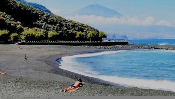 A beach with a rocky shoreline, a person lying on the sand, and a mountain in the background.
