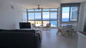 A spacious living area with a sofa, a TV, and a dining table with chairs. There is a large window offering a view of the sea. - Apartamento Piscinas Naturales (San Cristóbal de La Laguna)