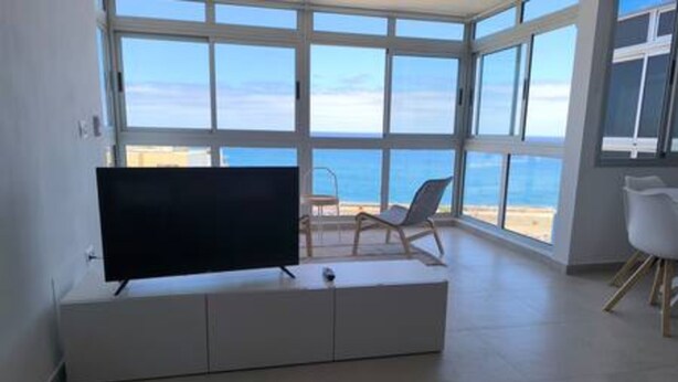 A room with a large window, a flat-screen TV on a white cabinet, and two white chairs. - Apartamento Piscinas Naturales (San Cristóbal de La Laguna)